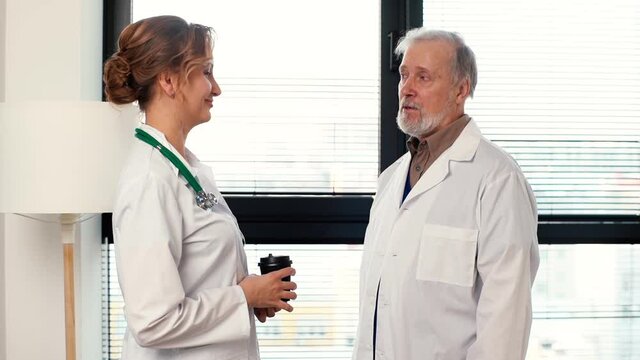 Woman doctor and mature adult physician surgeon having conversation, drinking coffee during break, standing near window at hospital. Male and female doctors discussing work and sipping hot drink.