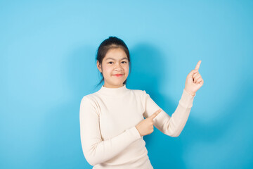 Portrait of smiling Asian teenager  joyfully and pointing finger to the side, Wears in white knitted sweater isolated over blue background with copy space