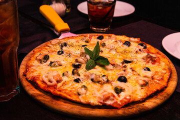An appetizing pizza with seafood and cheese lies on a wooden plate on the table. With a cold brown iced drink.