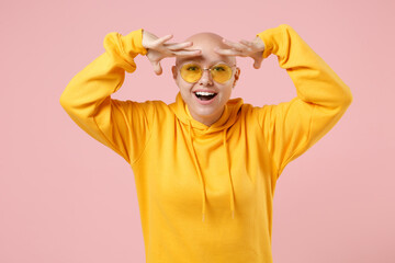 Young bald smiling caucasian woman 20s without hair wearing yellow casual sweatshirt shirt holding hand at forehead looking far away distance camera isolated on pastel pink background studio portrait.