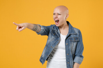 Young bald caucasian indignant strict serious woman 20s without hair in casual denim jacket white t-shirt point index finger aside screaming shouting isolated on yellow background studio portrait.