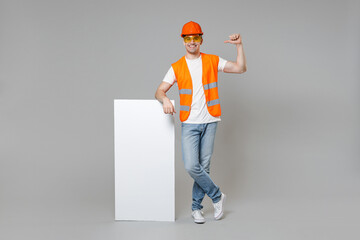 Full length employee man in orange vest hardhat hold blank sign board, place for text point finger on himself isolated on grey background studio. Instruments for renovation apartment. Repair concept.