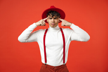Young spanish latinos pensive thoughtful stylish fashionable european man 20s in hat white shirt trousers, suspenders look camera keeps fingers on temples isolated on red background studio portrait.