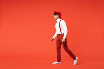 Full length of young spanish latinos attractive handsome smiling stylish fashionable european man 20s in hat white shirt trousers with suspenders walk going isolated on red background studio portrait.