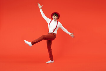Fototapeta na wymiar Full length of young spanish latinos surprised stylish man 20s in hat white shirt trousers with suspenders leaning over with outstratched hands raised up leg isolated on red background studio portrait