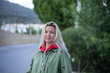 Closeup portrait of a beautiful forty-year-old blonde caucasian woman in a casual green jacket posing outdoors in a park against the background of green trees.