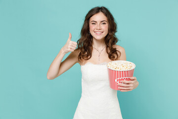 Smiling bride young woman 20s in beautiful white wedding dress hold bucket of popcorn showing thumb up like isolated on blue turquoise background studio portrait. Ceremony celebration party concept.