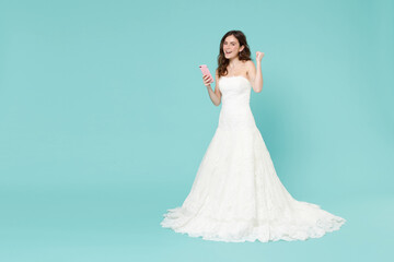 Full length of joyful happy bride young woman 20s in white wedding dress using mobile cell phone doing winner gesture say yes isolated on blue turquoise background. Ceremony celebration party concept.
