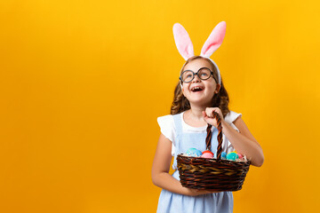 Cute little girl holding an Easter basket with eggs on a yellow background. Happy kid lifted his head and looks up. Copy space.