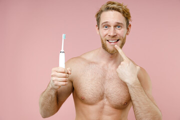 Smiling bearded naked young man 20s perfect skin hold electric brush pointing index finger on teeth...