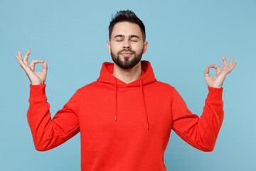 Fototapeta premium Young caucasian bearded man 20s in casual red orange hoodie hold hands in yoga gesture, relaxing meditating, trying to calm down isolated on blue background studio portrait People lifestyle concept.