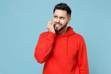Fototapeta premium Young caucasian troubled pensive stressed worried bearded man 20s in casual red orange hoodie looking aside biting nails finger isolated on blue background studio portrait People lifestyle concept.