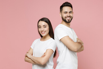 Young cheerful confident couple two friends man woman in white basic blank t-shirt standing bacl to back with folded crossed hands look camera isolated on pastel pink color background studio portrait
