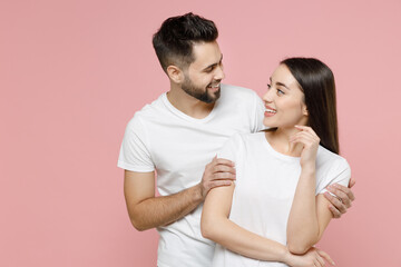 Young happy couple in love two friends man brunette woman 20s in white basic t-shirts looking to each other standing smiling hugging embrace isolated on pastel pink color background studio portrait.