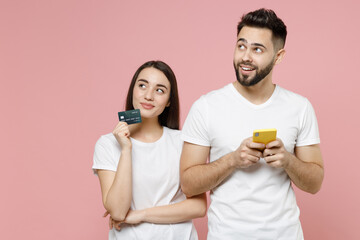 Young couple two friends man woman in white basic blank print design tshirts hold in hand using mobile cell phone credit bank card order home delivery in restaurant isolated on pastel pink background