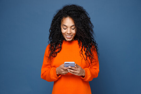 Smiling beautiful young african american woman wearing casual basic bright orange sweatshirt standing using mobile cell phone typing sms message isolated on blue color background studio portrait.
