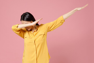 Young brunette cute stylish fashionable woman 20s in yellow shirt doing dab hip hop dance hands move gesture youth sign hiding covering face isolated on pastel pink color background studio portrait.