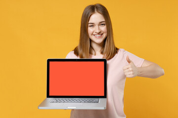 Naklejka premium Young freelancer woman 20s in casual basic pastel pink t-shirt holding laptop pc computer with blank screen workspace area show thumb up like gesture isolated on yellow background studio portrait