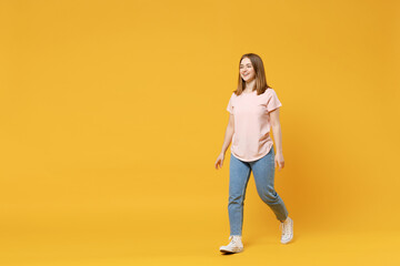 Full length of young friendly smiling student woman 20s with nude make up wearing casual basic pastel pink t-shirt, jeans looking aside, walking going isolated on yellow background studio portrait.