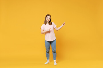 Full length of young caucasian woman 20s wear casual basic pastel pink t-shirt, jeans point index finger aside on workspace area mock up copy space isolated on yellow color background studio portrait.