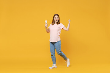 Full length of young caucasian student woman 20s in basic pastel pink t-shirt, jeans holding mobile cell phone do winner gesture clench fist celebrating isolated on yellow background studio portrait.