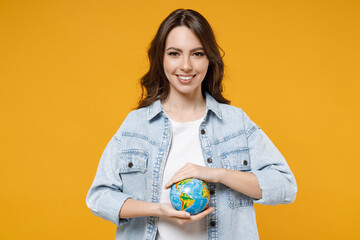 Young smiling happy brunette fun geography student woman 20s wearing stylish casual denim shirt white t-shirt holding in palms Earth world globe isolated on yellow color background studio portrait