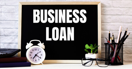 The word BUSINESS LOAN is written on the chalkboard next to the white alarm clock, glasses, potted...