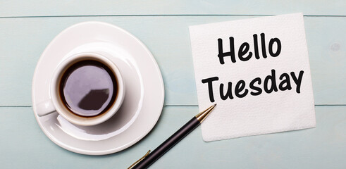 On a light blue wooden tray, there is a white cup of coffee, a handle and a napkin that says HELLO TUESDAY