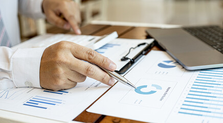 A close-up view of a businessman holding a pen pointing at a bar chart on a corporate finance document, he is meeting with senior management, with the theme of the meeting being Company Finance.