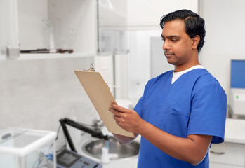 healthcare, profession and medicine concept - indian doctor or male nurse in blue uniform writing medical report on clipboard over laboratory or hospital background