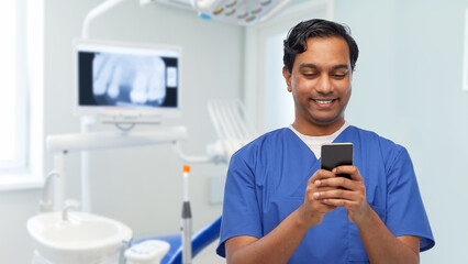 medicine, dentistry and technology concept - happy smiling indian doctor or male dentist in blue uniform using smartphone over dental clinic background