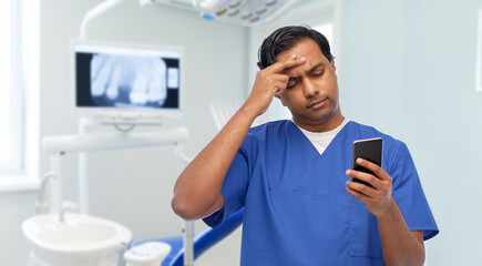 medicine, dentistry and technology concept - indian doctor or male dentist in blue uniform using smartphone over dental clinic background