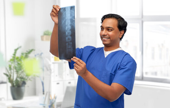 medicine, radiology and healthcare concept - happy smiling indian doctor or male nurse in blue uniform looking at x-ray scan image of spine over medical office at hospital background