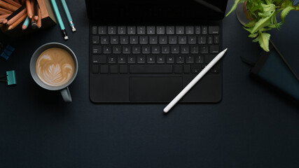 Overhead shot of wireless keyboard, stylus pen, coffee cup and plant on modern dark workplace.
