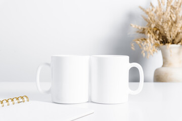 Two mugs mockup with workspace accessories, dried flowers in vase and notebook on white table. Front view. Place for text, copy space, mockup