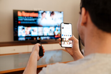 Caucasian guy using mobile phone while watching TV at home