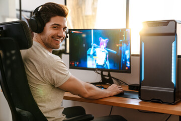 Happy brunette guy in headphones playing video game on his computer