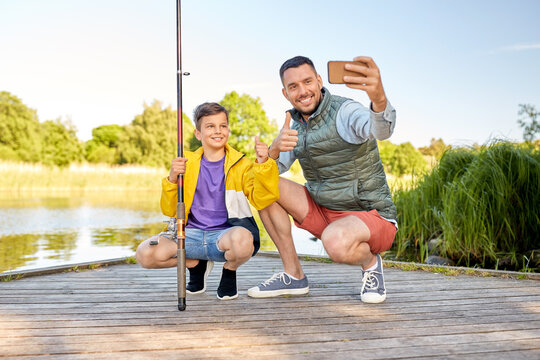 family, generation, summer holidays and people concept - happy smiling father and little son with fishing rods taking selfie with smartphone and showing thumbs up on river