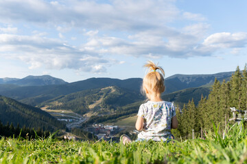 Fototapeta na wymiar Cute little young child sitting at green grass at the top of the hill with the view of mountains at summer, blue sky with clouds, view from the back.