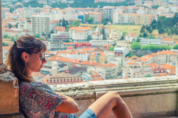 Young beautiful woman traveler with sunglasses is crossing arms and sitting on balcony of medieval Leiria castle in Portugal, old town and residential districts background