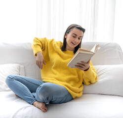 A cute girl in a yellow sweater is reading a book at home on the couch.