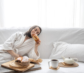 Young woman in white robe at home on the couch enjoying pastries and tea copy space.