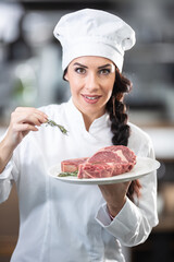 Beautiful female chef in uniform and hat holds a plate with raw steaks and fresh herbs in the kitchen