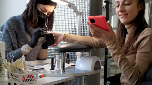 Professional blogger types on smartphone with nose and shows photos with smile while beautician makes manicure. Concept fun
