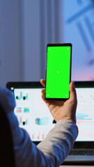 Woman employee looking at smartphone with chroma key sitting at desk in start up business office late at night. Business woman watching desktop display with green mockup, screen, working overtime.