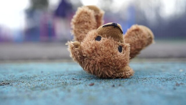 Lonely Teddy bear with sad face lying on footpath with blurry kids playing in playground at public park in gloomy day, Lost toy or Loneliness concept, International missing Children