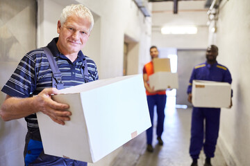 Older warehouse worker carries cardboard boxes through aisle