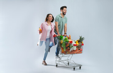 Young couple with shopping cart full of groceries on grey background