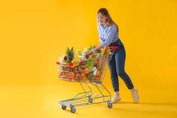Young woman with shopping cart full of groceries on yellow background