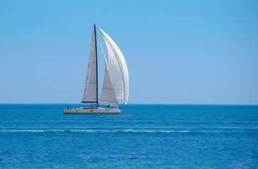 Sailboat with white sails on the open sea against the backdrop of a clear blue sky.